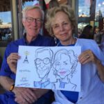 An older man and woman sit together, smiling and holding a Caricatures Bastille Day Healdsburg portrait of themselves.