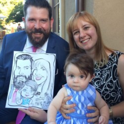 A man and woman with a baby pose together, holding a caricature drawing of themselves at an outdoor event.