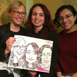 Three women smile while holding a caricature drawing of themselves, seated together in a warmly lit room.