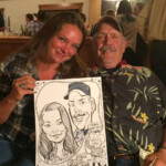 A woman and a man sit together indoors, smiling and holding a caricature drawing of themselves at an event.