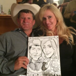 A man and woman sit together holding a caricature portrait of themselves at an indoor event.