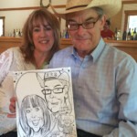 A smiling couple poses together while the man holds a caricature drawing of both of them at an indoor event.