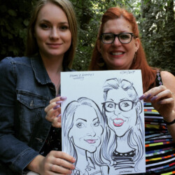 Two women sit outdoors, holding up a caricature drawing of themselves, and smiling at the camera.