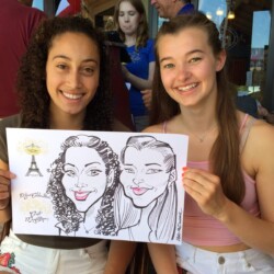 Two young women smile and hold a caricature drawing of themselves while sitting outdoors at a café.