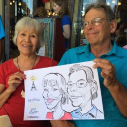 An older couple smiles while holding a caricature portrait of themselves at an outdoor café.