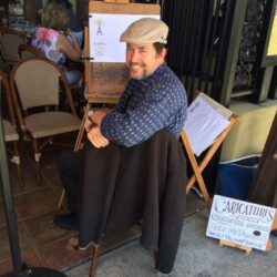 A man sits on a chair next to an easel with artwork and a sign advertising caricatures outside a café.