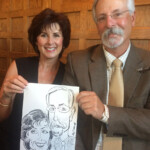 A smiling couple sits together, holding a caricature drawing of themselves against a wooden-paneled background.