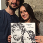 A man and woman smile and hold a caricature drawing of themselves against a textured wall.
