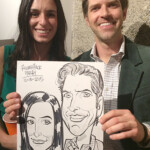 Two people smiling and holding a black-and-white caricature portrait of themselves at an event.