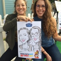 Two women smiling and holding a caricature drawing of themselves at an outdoor event.