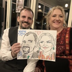 A man and woman smile while holding a caricature drawing of themselves at an indoor event labeled "Wedding Day.