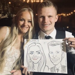 A smiling couple in formal attire holds a black-and-white caricature drawing of themselves at an outdoor event.