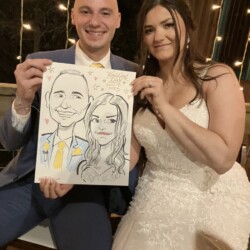 A bride and groom in wedding attire hold up a caricature drawing of themselves while sitting together.