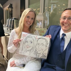 A woman in a white dress and a man in a suit smile while holding a caricature drawing of themselves at an event.