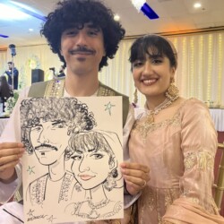 A smiling couple sits together at an event, holding a black-and-white caricature portrait of themselves.