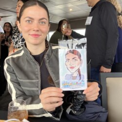 A woman sits at a table holding a caricature portrait of herself at an indoor social event.