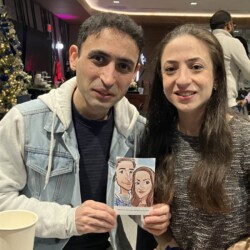 A man and woman sit indoors, holding a caricature drawing of themselves, with a Christmas tree in the background.