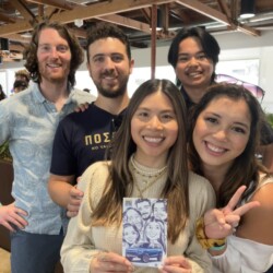 Five people smiling indoors, with one woman holding a caricature drawing of the group; wood beams visible above.