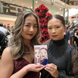 Two women pose indoors, holding a drawing of themselves in front of a large poinsettia Christmas tree.