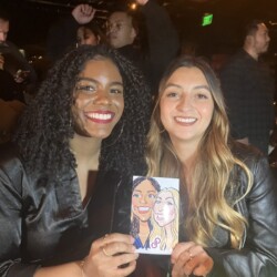 Two women smiling and holding a caricature drawing of themselves at an indoor event with people in the background.