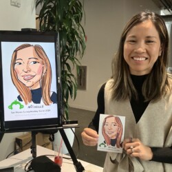 A woman smiles while holding a caricature portrait of herself, with the same drawing displayed on a screen next to her.